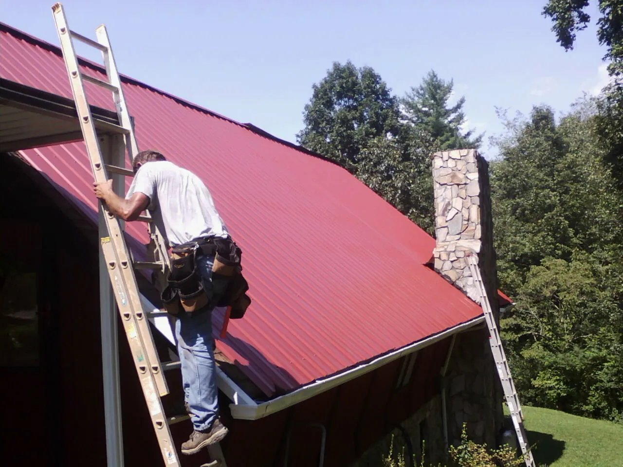 Roofer on a ladder beside a red metal roof and stone chimney — Soffit & Fascia Repair by Roofing Brothers in Winton